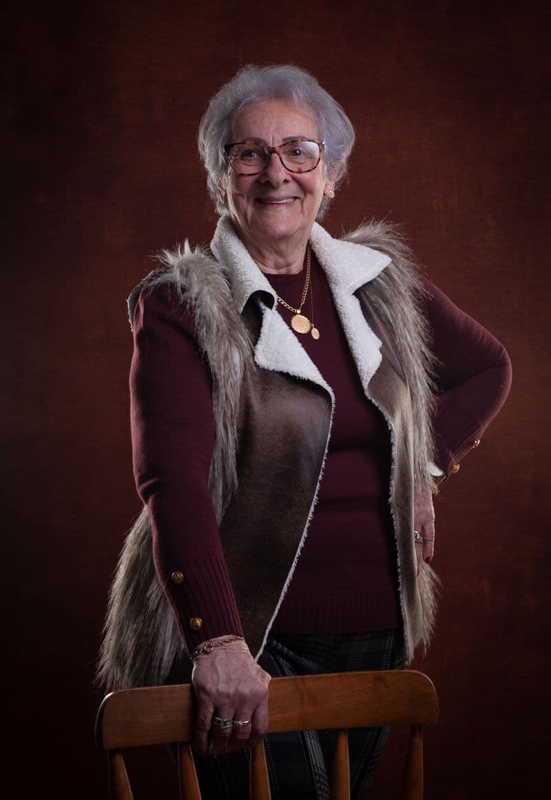 Ältere Dame mit Brille und Fellweste lehnt lächelnd an einer Holzstuhllehne